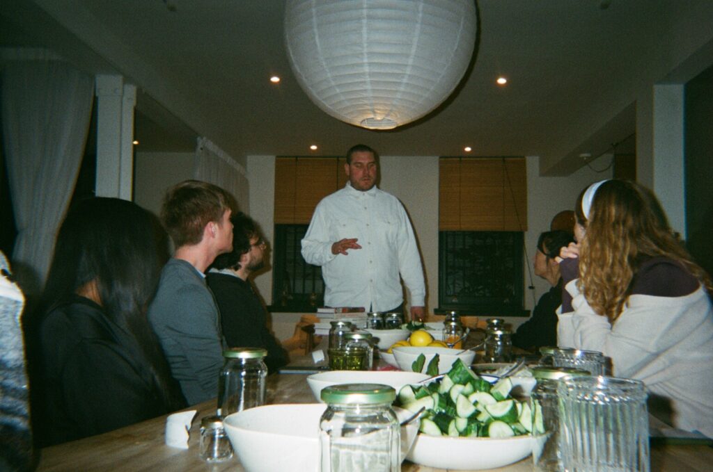 Six people gathered around a table looking at a chef explaining about pickling and fermentation. There are ingredients in the forefront of the image