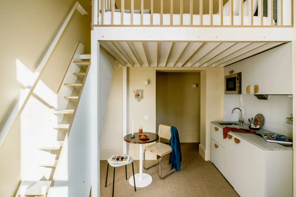 A wide image of Mason & Fifth, Primrose Hill's Mezzanine Studio of the wooden slatted stairs, mattress and kitchenette with a small table,wicker dining chair and stool.