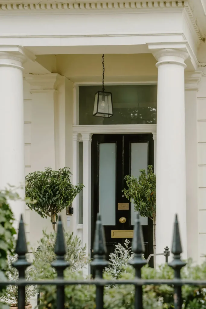 The black door and white porch pillars of Mason & Fifth, Primrose Hill, in the forefront a black iron fence and greenery.