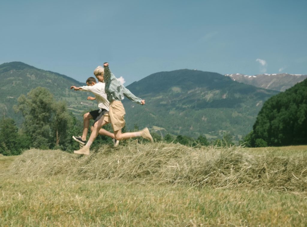 Two people jumping over a hay bale in a field with hilltops as the backdrop.